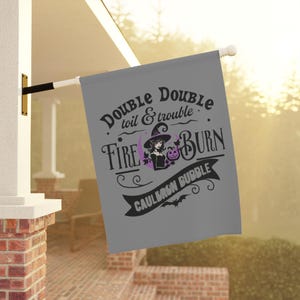 May include: A gray Halloween flag with the text "Double, double toil & trouble, fire burn, cauldron bubble." A witch illustration with a purple hat and a pumpkin is in the center. The flag is hanging outside.