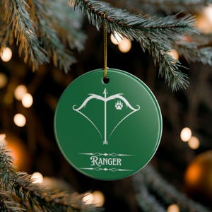 Op de afbeelding: Een ronde, groene kerstversiering met een wit pijl-en-boogontwerp, een pootafdruk en het woord "Ranger" in het wit. De versiering hangt aan een gouden draad tegen een vage achtergrond van kerstverlichting en een dennentak.