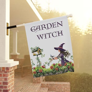 May include: A white garden flag with the words "GARDEN WITCH" in purple script. The flag features a whimsical illustration of a woman in a witch hat, a birdhouse, and flowers. The flag is hanging outside on a sunny day.