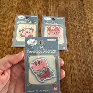 Könnte beinhalten: Drei Kirby Horoscope Collection Aufnäher. Jeder Aufnäher zeigt einen rosa Kirby-Charakter mit verschiedenen Designs, vor einem hellen Hintergrund. Die Aufnäher sind in durchsichtigem Kunststoff mit blau-weißem Text und Sterndesigns verpackt.