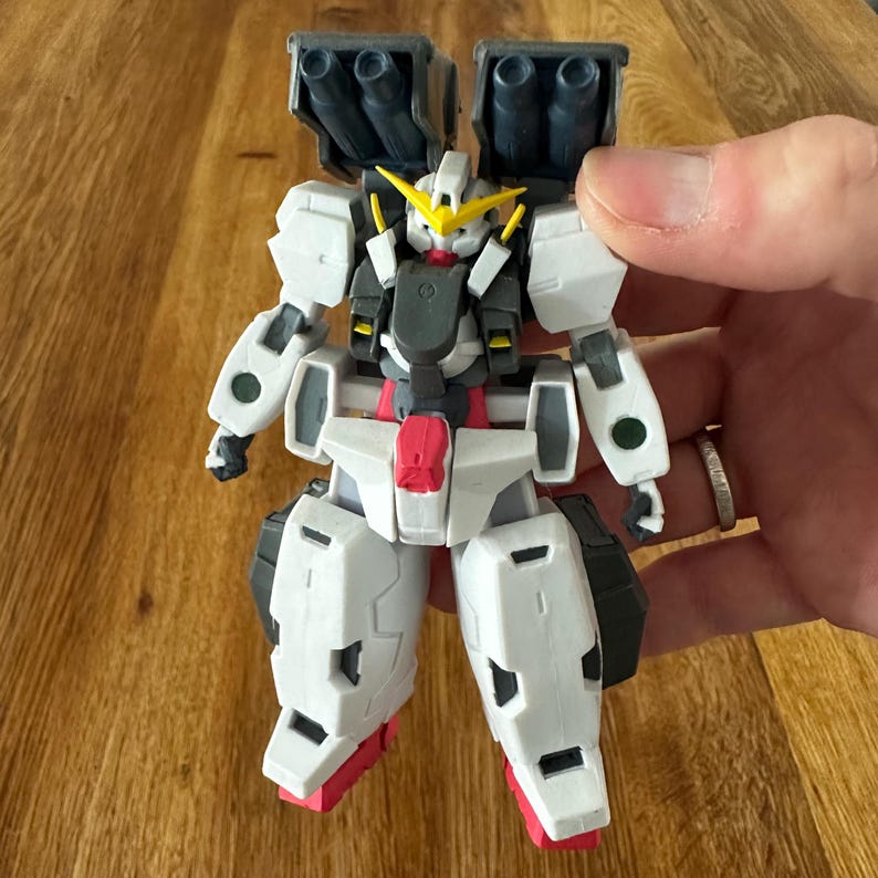 A close-up view of a completed Gundam Virtue (GN-005) action figure. The heavy assault mobile suit is made of high-quality plastic with a dominant white and grey color scheme. Dark grey cannons are mounted on its broad shoulders, and bright yellow fins extend from its head. The figure features black and red accents, including a red cockpit hatch and foot coverings. It is shown from multiple angles to highlight all armor panels, boosters, and joint details. The model is in excellent condition