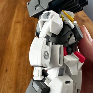 A close-up view of a completed Gundam Virtue (GN-005) action figure. The heavy assault mobile suit is made of high-quality plastic with a dominant white and grey color scheme. Dark grey cannons are mounted on its broad shoulders, and bright yellow fins extend from its head. The figure features black and red accents, including a red cockpit hatch and foot coverings. It is shown from multiple angles to highlight all armor panels, boosters, and joint details. The model is in excellent condition