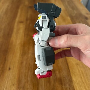 A close-up view of a completed Gundam Virtue (GN-005) action figure. The heavy assault mobile suit is made of high-quality plastic with a dominant white and grey color scheme. Dark grey cannons are mounted on its broad shoulders, and bright yellow fins extend from its head. The figure features black and red accents, including a red cockpit hatch and foot coverings. It is shown from multiple angles to highlight all armor panels, boosters, and joint details. The model is in excellent condition