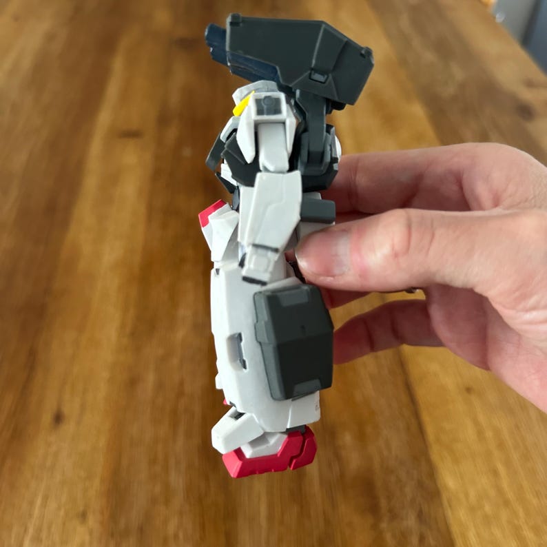 A close-up view of a completed Gundam Virtue (GN-005) action figure. The heavy assault mobile suit is made of high-quality plastic with a dominant white and grey color scheme. Dark grey cannons are mounted on its broad shoulders, and bright yellow fins extend from its head. The figure features black and red accents, including a red cockpit hatch and foot coverings. It is shown from multiple angles to highlight all armor panels, boosters, and joint details. The model is in excellent condition