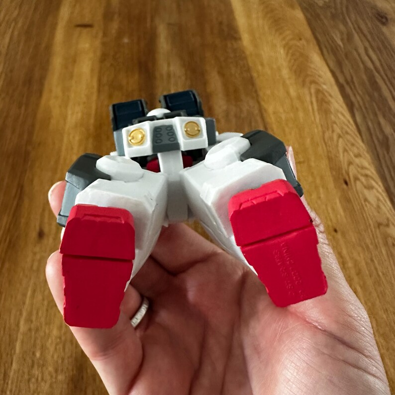 A close-up view of a completed Gundam Virtue (GN-005) action figure. The heavy assault mobile suit is made of high-quality plastic with a dominant white and grey color scheme. Dark grey cannons are mounted on its broad shoulders, and bright yellow fins extend from its head. The figure features black and red accents, including a red cockpit hatch and foot coverings. It is shown from multiple angles to highlight all armor panels, boosters, and joint details. The model is in excellent condition