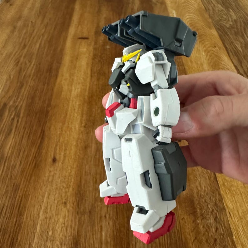 A close-up view of a completed Gundam Virtue (GN-005) action figure. The heavy assault mobile suit is made of high-quality plastic with a dominant white and grey color scheme. Dark grey cannons are mounted on its broad shoulders, and bright yellow fins extend from its head. The figure features black and red accents, including a red cockpit hatch and foot coverings. It is shown from multiple angles to highlight all armor panels, boosters, and joint details. The model is in excellent condition