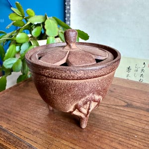 Puede incluir: Un quemador de incienso de cerámica marrón con tapa. El quemador tiene una superficie texturizada y tres patas. La tapa tiene un diseño floral y un pequeño asa. El quemador de incienso está sobre una superficie de madera.