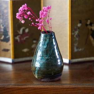 Può includere: Un piccolo vaso in ceramica affusolato con una smaltatura lucida e maculata nei toni del blu ottanio profondo, verde bosco e marrone scuro. Il vaso presenta un collo stretto e una base arrotondata più ampia, contenente un rametto di fiori rosa.