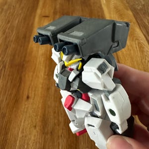 A pre-assembled Gundam Virtue GN-005 action figure from the 2007/2008 Gundam 00 anime series. The model features heavy white and grey plating with black and red accents. The large twin GN cannons are mounted on the shoulders. Multiple photos show the figure from different angles, including the back thrusters and side profile. It is shown held by hand to indicate scale, resting on a wooden background, and is in excellent, clean vintage condition.