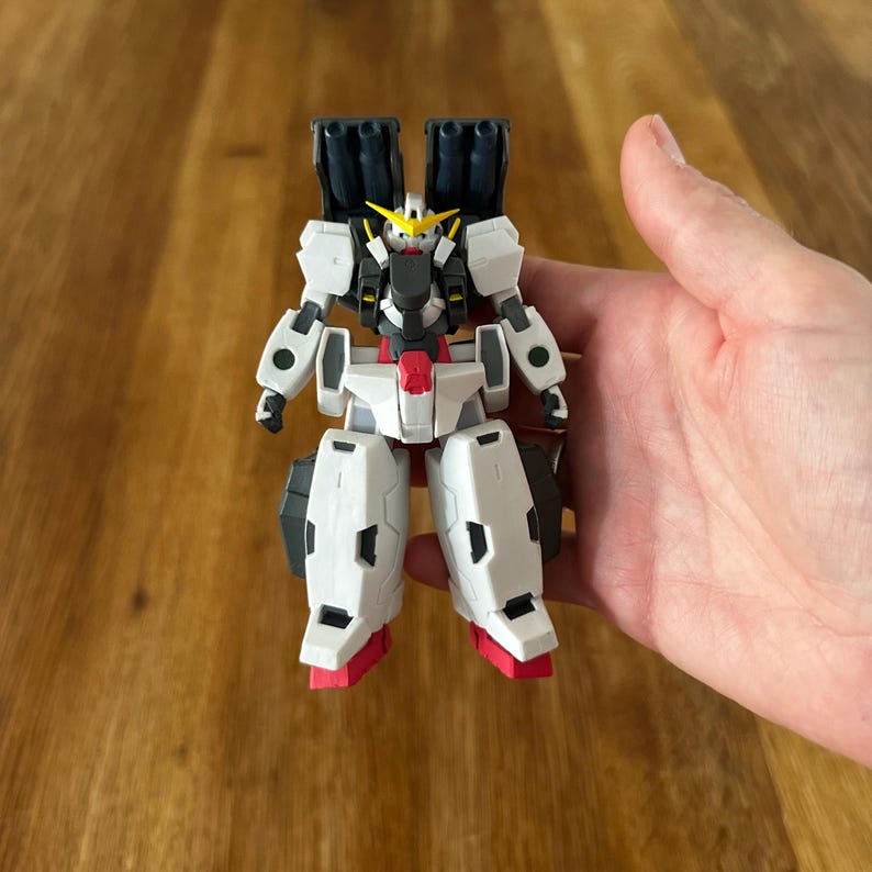 A close-up view of a completed Gundam Virtue (GN-005) action figure. The heavy assault mobile suit is made of high-quality plastic with a dominant white and grey color scheme. Dark grey cannons are mounted on its broad shoulders, and bright yellow fins extend from its head. The figure features black and red accents, including a red cockpit hatch and foot coverings. It is shown from multiple angles to highlight all armor panels, boosters, and joint details. The model is in excellent condition