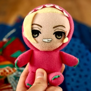 May include: A handmade plush doll in a pink hooded outfit with a smiling face. The doll has blonde hair, black eyes, and green accents on the feet and hands. The hood is trimmed with white and yellow details.