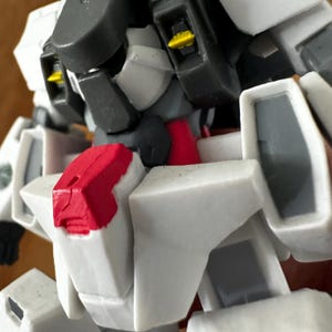 A close-up view of a completed Gundam Virtue (GN-005) action figure. The heavy assault mobile suit is made of high-quality plastic with a dominant white and grey color scheme. Dark grey cannons are mounted on its broad shoulders, and bright yellow fins extend from its head. The figure features black and red accents, including a red cockpit hatch and foot coverings. It is shown from multiple angles to highlight all armor panels, boosters, and joint details. The model is in excellent condition
