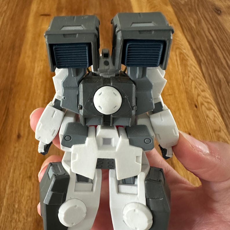 A close-up view of a completed Gundam Virtue (GN-005) action figure. The heavy assault mobile suit is made of high-quality plastic with a dominant white and grey color scheme. Dark grey cannons are mounted on its broad shoulders, and bright yellow fins extend from its head. The figure features black and red accents, including a red cockpit hatch and foot coverings. It is shown from multiple angles to highlight all armor panels, boosters, and joint details. The model is in excellent condition