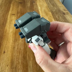 A close-up view of a completed Gundam Virtue (GN-005) action figure. The heavy assault mobile suit is made of high-quality plastic with a dominant white and grey color scheme. Dark grey cannons are mounted on its broad shoulders, and bright yellow fins extend from its head. The figure features black and red accents, including a red cockpit hatch and foot coverings. It is shown from multiple angles to highlight all armor panels, boosters, and joint details. The model is in excellent condition