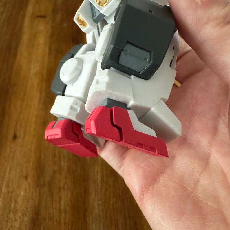 A close-up view of a completed Gundam Virtue (GN-005) action figure. The heavy assault mobile suit is made of high-quality plastic with a dominant white and grey color scheme. Dark grey cannons are mounted on its broad shoulders, and bright yellow fins extend from its head. The figure features black and red accents, including a red cockpit hatch and foot coverings. It is shown from multiple angles to highlight all armor panels, boosters, and joint details. The model is in excellent condition