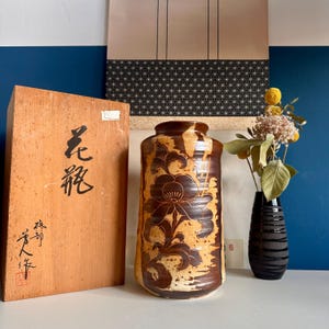 May include: A brown and tan ceramic vase with a floral design. The vase is next to a wooden box with Japanese characters and a black vase with flowers. The background includes a blue wall and a decorative scroll.