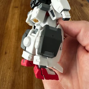 A close-up view of a completed Gundam Virtue (GN-005) action figure. The heavy assault mobile suit is made of high-quality plastic with a dominant white and grey color scheme. Dark grey cannons are mounted on its broad shoulders, and bright yellow fins extend from its head. The figure features black and red accents, including a red cockpit hatch and foot coverings. It is shown from multiple angles to highlight all armor panels, boosters, and joint details. The model is in excellent condition
