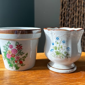 Può includere: Due vasi di fiori in ceramica bianca con bordi marroni. Il vaso più grande ha un motivo floreale con fiori rosa, rossi e verdi. Il vaso più piccolo ha un motivo floreale con fiori blu e verdi.