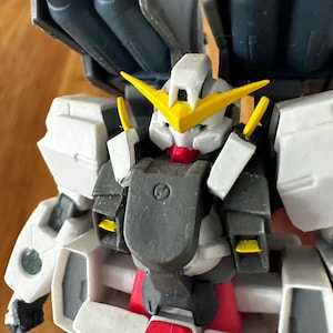 A close-up view of a completed Gundam Virtue (GN-005) action figure. The heavy assault mobile suit is made of high-quality plastic with a dominant white and grey color scheme. Dark grey cannons are mounted on its broad shoulders, and bright yellow fins extend from its head. The figure features black and red accents, including a red cockpit hatch and foot coverings. It is shown from multiple angles to highlight all armor panels, boosters, and joint details. The model is in excellent condition