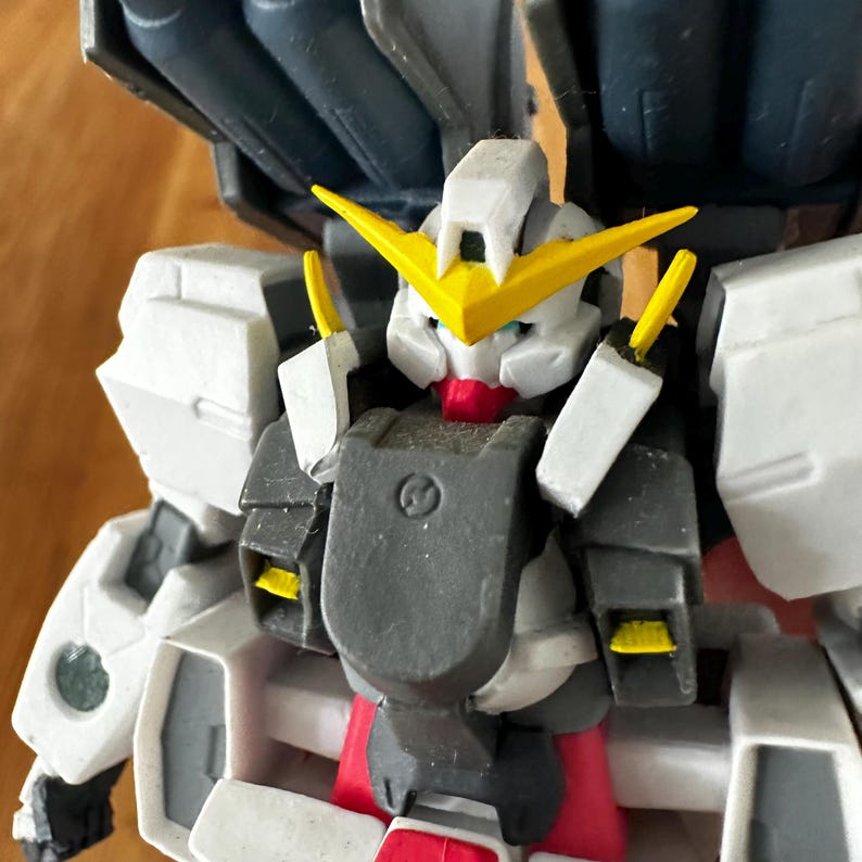 A close-up view of a completed Gundam Virtue (GN-005) action figure. The heavy assault mobile suit is made of high-quality plastic with a dominant white and grey color scheme. Dark grey cannons are mounted on its broad shoulders, and bright yellow fins extend from its head. The figure features black and red accents, including a red cockpit hatch and foot coverings. It is shown from multiple angles to highlight all armor panels, boosters, and joint details. The model is in excellent condition