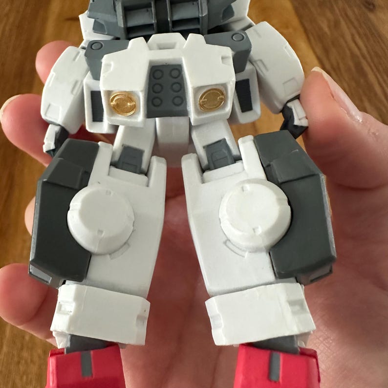 A close-up view of a completed Gundam Virtue (GN-005) action figure. The heavy assault mobile suit is made of high-quality plastic with a dominant white and grey color scheme. Dark grey cannons are mounted on its broad shoulders, and bright yellow fins extend from its head. The figure features black and red accents, including a red cockpit hatch and foot coverings. It is shown from multiple angles to highlight all armor panels, boosters, and joint details. The model is in excellent condition