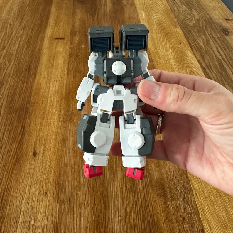 A close-up view of a completed Gundam Virtue (GN-005) action figure. The heavy assault mobile suit is made of high-quality plastic with a dominant white and grey color scheme. Dark grey cannons are mounted on its broad shoulders, and bright yellow fins extend from its head. The figure features black and red accents, including a red cockpit hatch and foot coverings. It is shown from multiple angles to highlight all armor panels, boosters, and joint details. The model is in excellent condition