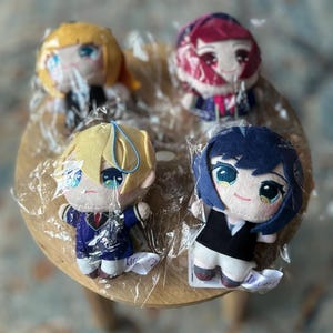 Może przedstawiać: Cztery małe pluszowe lalki z cechami w stylu anime, każda z unikalną fryzurą i strojem. Lalki są zapakowane w przezroczysty plastik i leżą na drewnianej powierzchni. Lalki mają duże oczy.