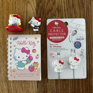 May include: A collection of Hello Kitty themed items, including figurines, a spiral-bound notebook, and two white iPhone cable protectors. The notebook features a Hello Kitty illustration with sweets. The cable protectors have Hello Kitty faces.