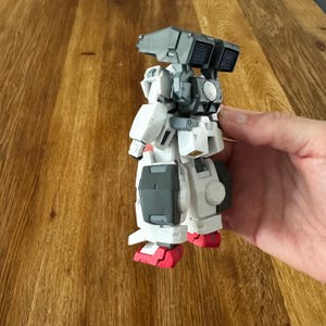 A close-up view of a completed Gundam Virtue (GN-005) action figure. The heavy assault mobile suit is made of high-quality plastic with a dominant white and grey color scheme. Dark grey cannons are mounted on its broad shoulders, and bright yellow fins extend from its head. The figure features black and red accents, including a red cockpit hatch and foot coverings. It is shown from multiple angles to highlight all armor panels, boosters, and joint details. The model is in excellent condition