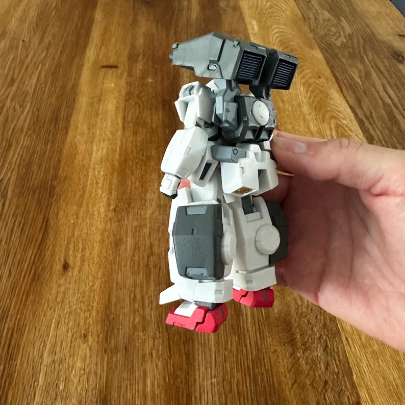 A close-up view of a completed Gundam Virtue (GN-005) action figure. The heavy assault mobile suit is made of high-quality plastic with a dominant white and grey color scheme. Dark grey cannons are mounted on its broad shoulders, and bright yellow fins extend from its head. The figure features black and red accents, including a red cockpit hatch and foot coverings. It is shown from multiple angles to highlight all armor panels, boosters, and joint details. The model is in excellent condition