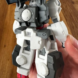 A close-up view of a completed Gundam Virtue (GN-005) action figure. The heavy assault mobile suit is made of high-quality plastic with a dominant white and grey color scheme. Dark grey cannons are mounted on its broad shoulders, and bright yellow fins extend from its head. The figure features black and red accents, including a red cockpit hatch and foot coverings. It is shown from multiple angles to highlight all armor panels, boosters, and joint details. The model is in excellent condition
