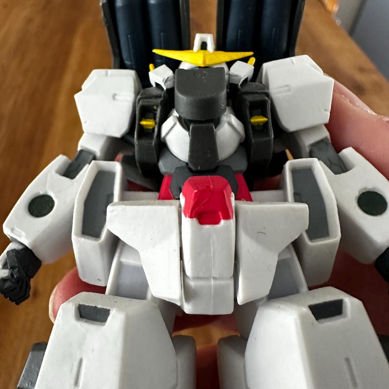A close-up view of a completed Gundam Virtue (GN-005) action figure. The heavy assault mobile suit is made of high-quality plastic with a dominant white and grey color scheme. Dark grey cannons are mounted on its broad shoulders, and bright yellow fins extend from its head. The figure features black and red accents, including a red cockpit hatch and foot coverings. It is shown from multiple angles to highlight all armor panels, boosters, and joint details. The model is in excellent condition