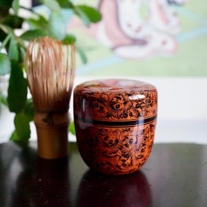 Può includere: Un contenitore per tè laccato arancione e nero con un motivo floreale. Un frullatore per matcha in bambù è a sinistra del contenitore. Il contenitore è rotondo con un coperchio e una fascia nera al centro. Lo sfondo è sfocato con fogliame verde e un dipinto.