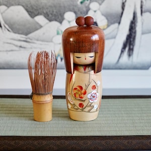 Pode incluir: Uma boneca Kokeshi japonesa de madeira com cabeça castanha e um desenho floral pintado no corpo. Ao lado, um batedor de matcha de bambu. A boneca tem cerca de 15 cm de altura.