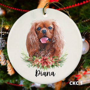 May include: A white ceramic Christmas ornament featuring a Cavalier King Charles Spaniel dog with brown fur, a pink tongue, and a floral wreath around its neck. The ornament is personalized with the name "Diana".