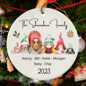 May include: White ceramic Christmas ornament with a gnome family illustration. The ornament features five gnomes, two cats, and the text "The Shanahan Family, Nancy - Bill - Katie - Morgan, Baby - Chip, 2023".