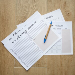 Puede incluir: Un conjunto de tres planificadores imprimibles blancos con texto negro. Los planificadores están etiquetados como "MON Planning JOURNALIER", "ANNUEL" y "MENSUEL". Un bolígrafo azul y marrón está sobre los planificadores.