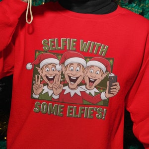 May include: A person wearing a red sweatshirt with the text "SELFIE WITH SOME ELFIE'S!" and a graphic of three cartoon elves. The person is holding a yellow camera in front of a Christmas tree.