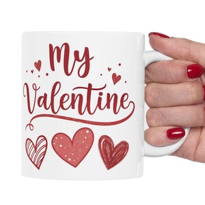 Può includere: Tazza in ceramica bianca con un design rosso e bianco che dice "My Valentine" con piccoli cuori intorno al testo e tre cuori più grandi in fondo.