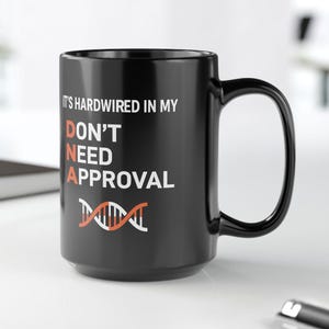 May include: A black ceramic mug with the text "IT'S HARDWIRED IN MY DON'T NEED APPROVAL" in white and orange. The mug features a DNA double helix graphic. The mug is on a white surface.