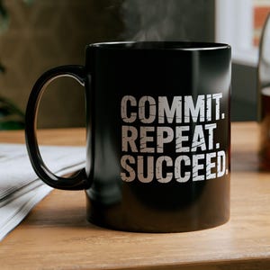 May include: A black mug with the text "COMMIT. REPEAT. SUCCEED" in white. Steam rises from the mug, which sits on a wooden surface. A glass carafe with a brown liquid is visible in the background.