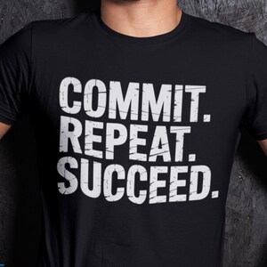 Puede incluir: Una camiseta negra de manga corta con el texto blanco "COMMIT. REPEAT. SUCCEED." impreso en una fuente desgastada en el pecho. La camiseta tiene cuello redondo y se lleva con vendas azules en las muñecas y pantalones cortos deportivos negros.