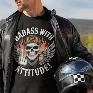 May include: Black t-shirt featuring a skull graphic with goggles, wings, and the text "BADASS WITH ATTITUDE!". The person is wearing a black leather jacket and holding a black motorcycle helmet.