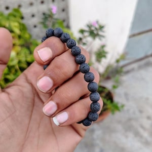 Puede incluir: Una pulsera elástica compuesta por numerosas cuentas esféricas pequeñas de piedra volcánica negra. Cada cuenta presenta una superficie porosa y texturizada característica de la roca volcánica. Las cuentas están unidas en un bucle continuo.