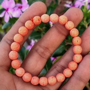 Può includere: Un braccialetto elastico composto da perline rotonde, porose e color pesca. Le perline presentano una superficie ruvida e bucherellata, tipica della roccia vulcanica. Il filo elastico arancione tiene unite le perline in un cerchio.