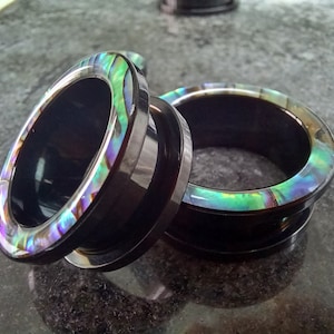 May include: Two black and iridescent ear gauges with a smooth, polished finish. The gauges have a wide, flat band with a thin, iridescent ring around the edge.