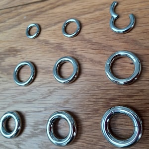May include: A collection of shiny, silver-colored body jewelry, including various sizes of circular rings and a hinged ring. The jewelry is arranged on a wooden surface, showcasing the polished metal and the different diameters available.