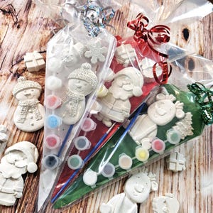 May include: Christmas-themed craft kits in clear cellophane bags. Each kit contains a plaster snowman, Santa, or reindeer figure, paint pots, and a paintbrush. The bags are tied with festive ribbons and bows, and include additional plaster shapes.