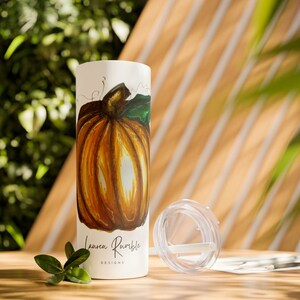May include: White tumbler with a hand-painted orange pumpkin design. The tumbler has a clear lid and a straw. The text "Lauren Rumble Designs" is printed on the bottom of the tumbler.