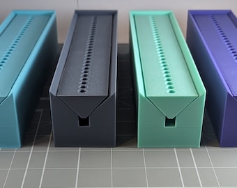 May include: Four rectangular, 3D-printed storage boxes in shades of blue, gray, green, and purple. Each box has a series of evenly spaced holes along the top and a square opening on the front. The boxes are arranged side by side.